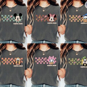 May include: Dark gray t-shirts featuring various Disney characters like Mickey Mouse, Minnie Mouse, Donald Duck, Pluto, Daisy Duck, and Goofy. Each shirt has a checkered pattern with flowers and the text "CUSTOM NAME".