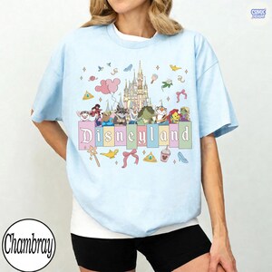Pu&ograve; includere: T-shirt azzurra con grafica colorata di Disneyland. Il design include il castello di Disneyland, vari personaggi dei cartoni animati e la scritta "Disneyland" in colori pastello. La maglietta ha maniche corte e girocollo.
