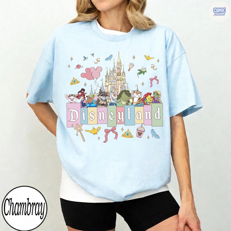 Pu&ograve; includere: T-shirt azzurra con grafica colorata di Disneyland. Il design include il castello di Disneyland, vari personaggi dei cartoni animati e la scritta "Disneyland" in colori pastello. La maglietta ha maniche corte e girocollo.