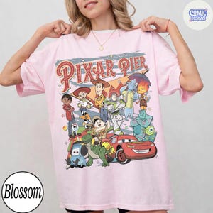 Pu&ograve; includere: Una maglietta rosa chiaro con una grafica colorata di vari personaggi Pixar, tra cui Woody, Buzz Lightyear e Saetta McQueen. Le parole "Pixar Pier" sono stampate sopra i personaggi in un font retr&ograve;. La maglietta ha maniche corte e una vestibilit&agrave; ampia.