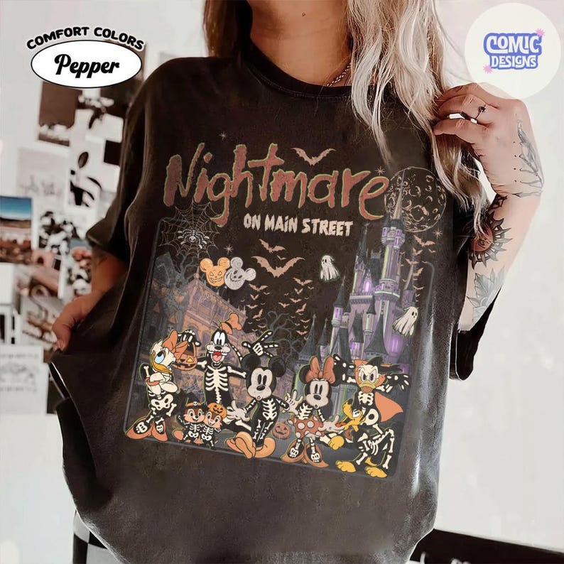 May include: A charcoal gray t-shirt featuring a graphic print with the text Nightmare ON MAIN STREET. The design includes cartoon characters dressed in skeleton costumes, a castle, bats, a spider web, and a full moon in a Halloween theme.