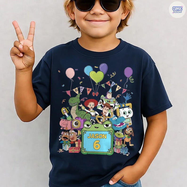 May include: A navy blue short-sleeve t-shirt featuring a colorful graphic of various animated toy characters, balloons, and a banner that reads BIRTHDAY. A central green frog-shaped frame displays the name JASON and the number 6 in yellow text.