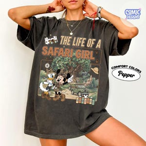May include: Dark gray t-shirt with a safari-themed graphic featuring Minnie Mouse and Daisy Duck. The text reads "THE LIFE OF A SAFARI GIRL" with a safari scene illustration. The shirt is from Comfort Colors.