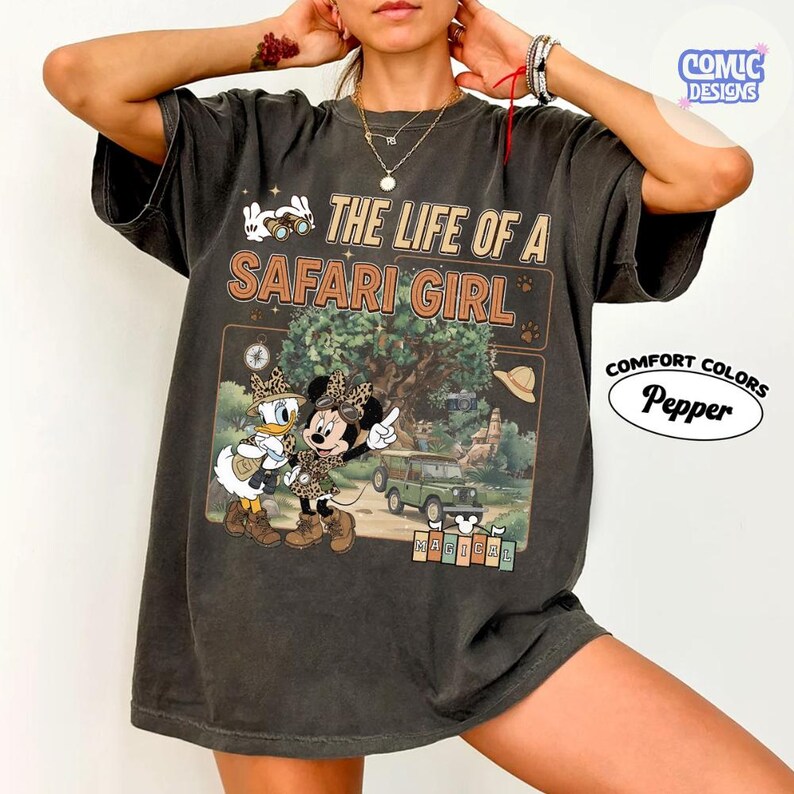 May include: Dark gray t-shirt with a safari-themed graphic featuring Minnie Mouse and Daisy Duck. The text reads "THE LIFE OF A SAFARI GIRL" with a safari scene illustration. The shirt is from Comfort Colors.