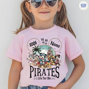 May include: Pink t-shirt featuring a pirate-themed design with cartoon characters. The design includes the word "PIRATES" in a bold font, the text "Life for Me", and the year "2026". The shirt has a crew neck and short sleeves.