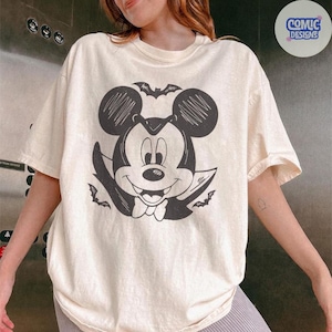 May include: Cream-colored t-shirt featuring a black and white graphic of Mickey Mouse dressed as a vampire, complete with bat wings and a cape. The design is centered on the front of the shirt. The shirt has short sleeves and a crew neck.