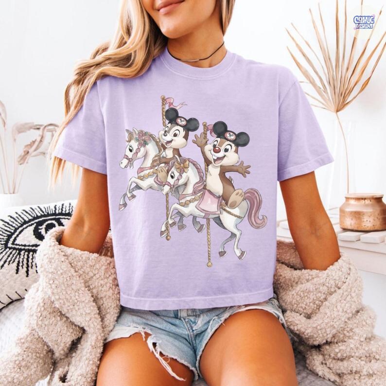 May include: A light purple t-shirt featuring a carousel scene with cartoon characters. The carousel horses are white with pink accents, and the characters are wearing hats. The shirt has a crew neck and short sleeves.