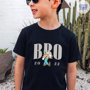 K&ouml;nnte beinhalten: Schwarzes T-Shirt mit dem Wort "BRO" in Grau, zusammen mit einer Cartoon-Charakter-Illustration und den Zahlen "20 22" darunter. Das T-Shirt wird von einer Person getragen.