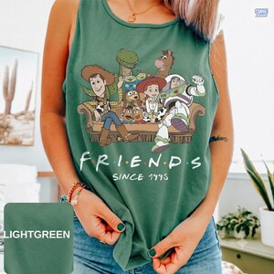 May include: A light green sleeveless tank top featuring a graphic print of various animated characters sitting on a couch. The text below the characters reads FRIENDS SINCE 1995. The design includes a cowboy, a space ranger, and other figures.