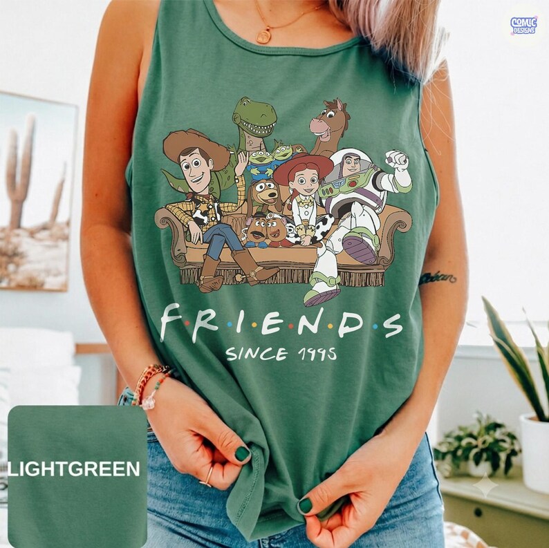 May include: A light green sleeveless tank top featuring a graphic print of various animated characters sitting on a couch. The text below the characters reads FRIENDS SINCE 1995. The design includes a cowboy, a space ranger, and other figures.