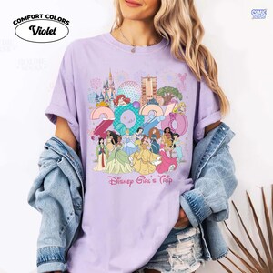 May include: A violet short-sleeved t-shirt featuring a graphic print of the year 2024, various Disney princesses, and theme park landmarks. The text Disney Girl's Trip is printed below the graphic. The shirt has a crew neckline and a relaxed fit.