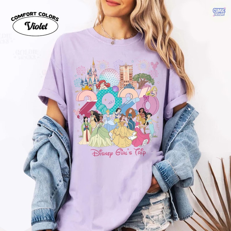 May include: A violet short-sleeved t-shirt featuring a graphic print of the year 2024, various Disney princesses, and theme park landmarks. The text Disney Girl's Trip is printed below the graphic. The shirt has a crew neckline and a relaxed fit.