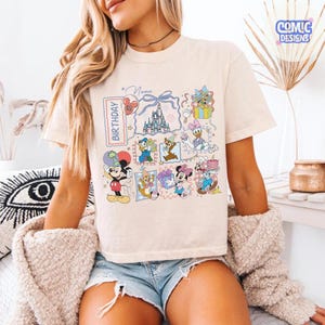 May include: Cream-colored t-shirt featuring a colorful graphic of Disney characters and a castle. The design includes Mickey Mouse, Minnie Mouse, Donald Duck, and Chip and Dale. The word "BIRTHDAY" is printed vertically on the left side of the graphic.