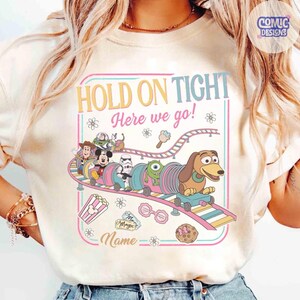 May include: Cream-colored t-shirt featuring a colorful amusement park design. The design includes the text "HOLD ON TIGHT Here we go!" and a train with cartoon characters. Additional graphics include popcorn, ice cream, and a cookie.
