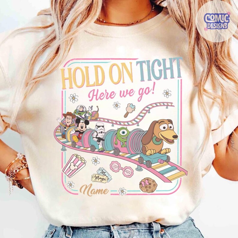 May include: Cream-colored t-shirt featuring a colorful amusement park design. The design includes the text "HOLD ON TIGHT Here we go!" and a train with cartoon characters. Additional graphics include popcorn, ice cream, and a cookie.