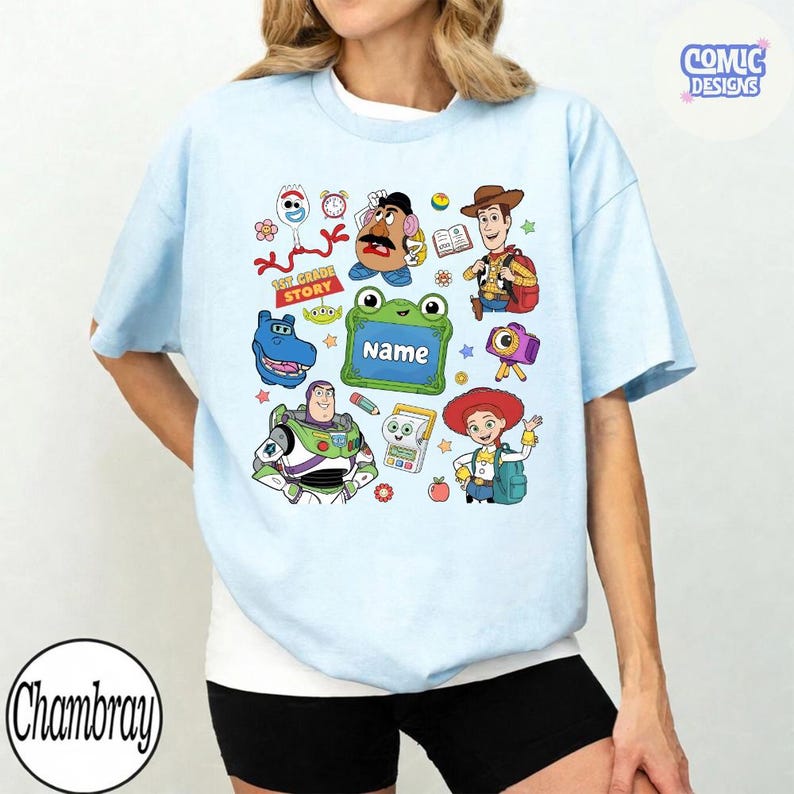 May include: A light blue t-shirt featuring a colorful graphic print of various Toy Story characters including Woody, Buzz Lightyear, Jessie, Mr. Potato Head, and Forky, along with a green frog-shaped frame with the text Name and 1st Grade Story.