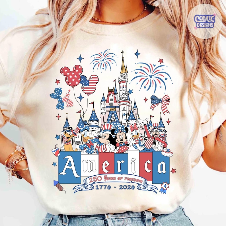 May include: A cream-colored t-shirt featuring a graphic of a castle, fireworks, and cartoon characters in patriotic red, white, and blue. The text reads America, 250 Years of Freedom, and 1776-2026, with Mickey-shaped balloons and stars.