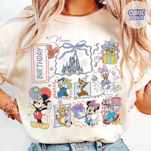 May include: Cream-colored t-shirt featuring a colorful birthday design with Disney characters. The design includes a castle, balloons, and the word "Birthday". Mickey and Minnie Mouse, Donald Duck, and other characters are also featured.