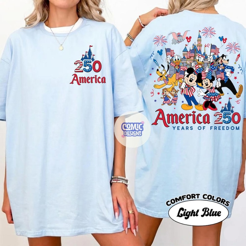 以下が含まれることがあります： 「250 America」の文字と城のイラストが描かれたライトブルーのTシャツ。Tシャツの背面には、漫画のキャラクター、花火、そして「America 250 Years of Freedom」の文字が描かれたカラフルなデザインが施されています。