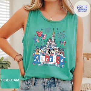 May include: A seafoam green sleeveless tank top featuring a graphic print of a castle, fireworks, and cartoon characters wearing patriotic red, white, and blue accessories. The text reads America, 250 Years of Freedom, 1776-2026 in a banner.
