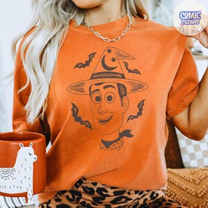 May include: Orange t-shirt featuring a Woody-inspired design with a witch hat, bats, and a smiling face. The shirt has a crew neck and short sleeves. A brown ceramic mug with a white llama design is also visible.