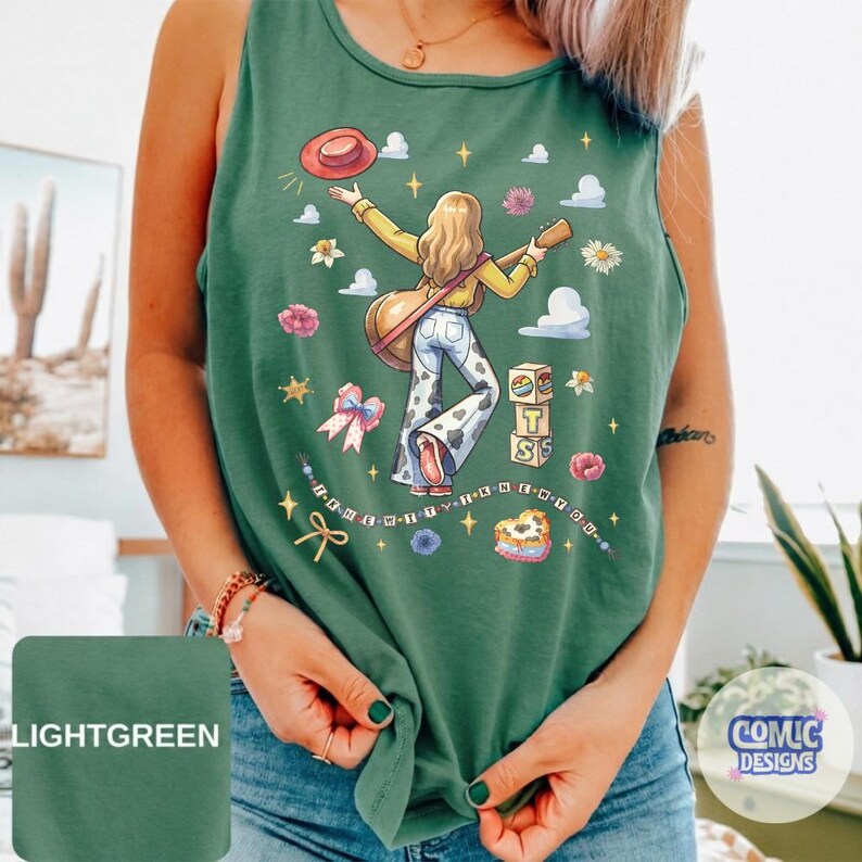 May include: A light green tank top featuring a graphic illustration of a person with long hair playing an acoustic guitar, wearing cow-print pants. The design includes a red hat, clouds, flowers, a bow, blocks with the letters T S, and text.