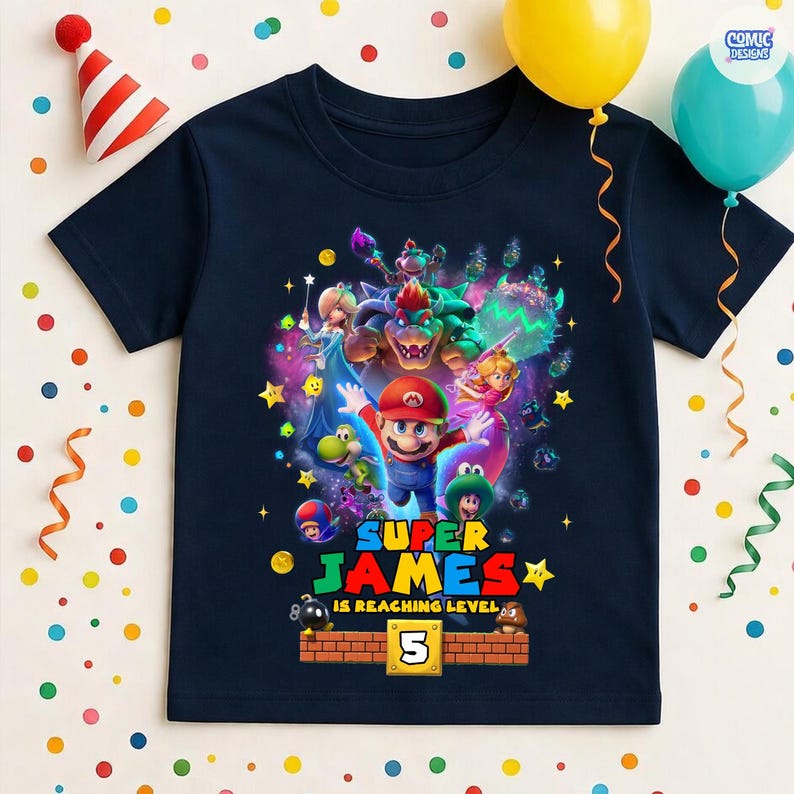 May include: Navy blue t-shirt featuring a colorful graphic of Super Mario characters. The design includes the text "SUPER JAMES IS REACHING LEVEL 5" with a brick-style border. Party decorations, including balloons and a party hat, are in the background.