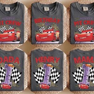 Puede incluir: Una colecci&oacute;n de seis camisetas gris oscuro con estampados gr&aacute;ficos de un coche de carreras rojo, banderas a cuadros, telara&ntilde;as y murci&eacute;lagos. Las camisetas muestran textos como Henry's Pit Crew, Birthday Boy, Dada, Henry y Mama, junto con el n&uacute;mero 1.