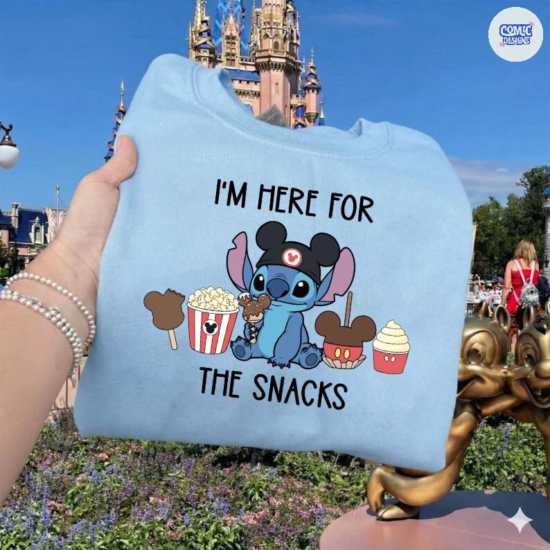 May include: Light blue sweatshirt featuring a graphic of the character Stitch wearing a Mickey Mouse hat, surrounded by snacks. The text "I'M HERE FOR THE SNACKS" is printed above and below the graphic. The sweatshirt is held up by a person.