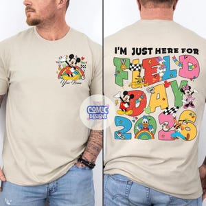 K&ouml;nnte beinhalten: Beiges T-Shirt mit Mickey Mouse auf einem Regenbogen und dem Text "Your Name" auf der Vorderseite. Auf der R&uuml;ckseite des Shirts steht "I'm just here for Field Day 2023" mit Disney-Figuren und bunten Grafiken.
