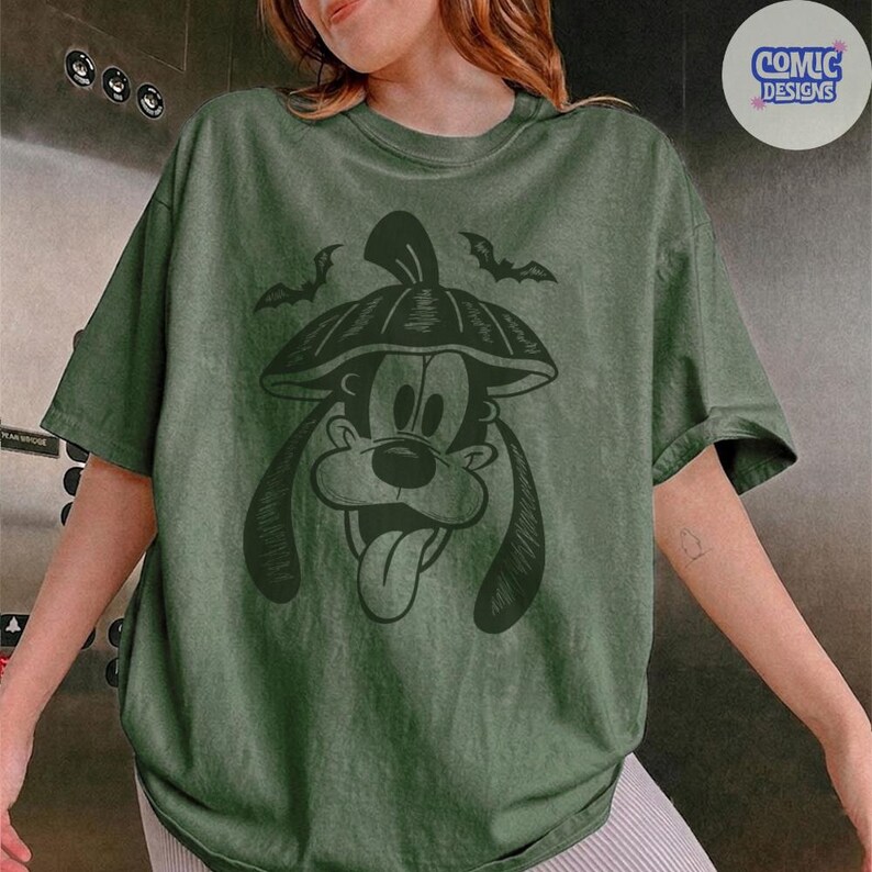 May include: Green t-shirt featuring a black graphic of a cartoon dog wearing a pumpkin hat, with two bats above. The shirt has a relaxed fit and a vintage wash appearance. The Comic Designs logo is in the upper right corner.
