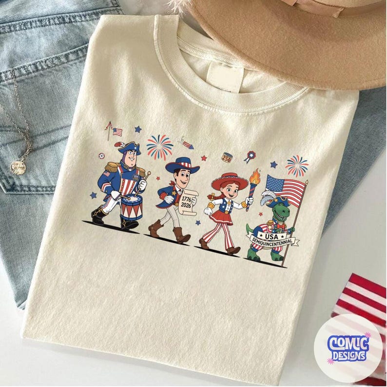 May include: Cream-colored t-shirt featuring a patriotic parade of cartoon characters. The design includes a drummer, a figure holding a scroll with "1776 2026", a torchbearer, and a dinosaur with a flag. The shirt is decorated with fireworks and stars.