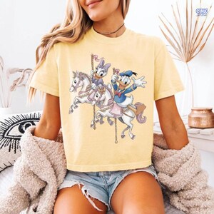 May include: Yellow t-shirt featuring a carousel scene with Donald Duck and Daisy Duck riding white horses. The graphic has a cartoon style with pastel colors. The shirt has short sleeves and a crew neck.