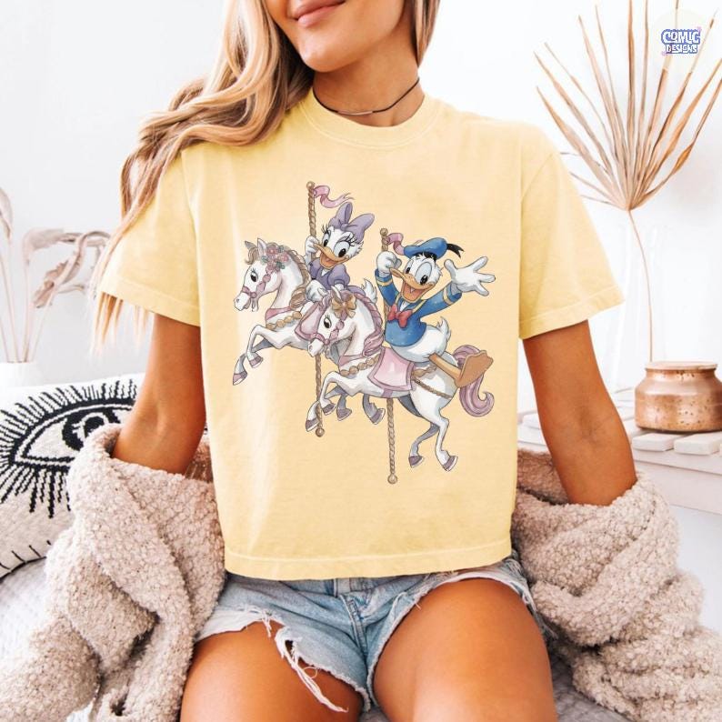 May include: Yellow t-shirt featuring a carousel scene with Donald Duck and Daisy Duck riding white horses. The graphic has a cartoon style with pastel colors. The shirt has short sleeves and a crew neck.