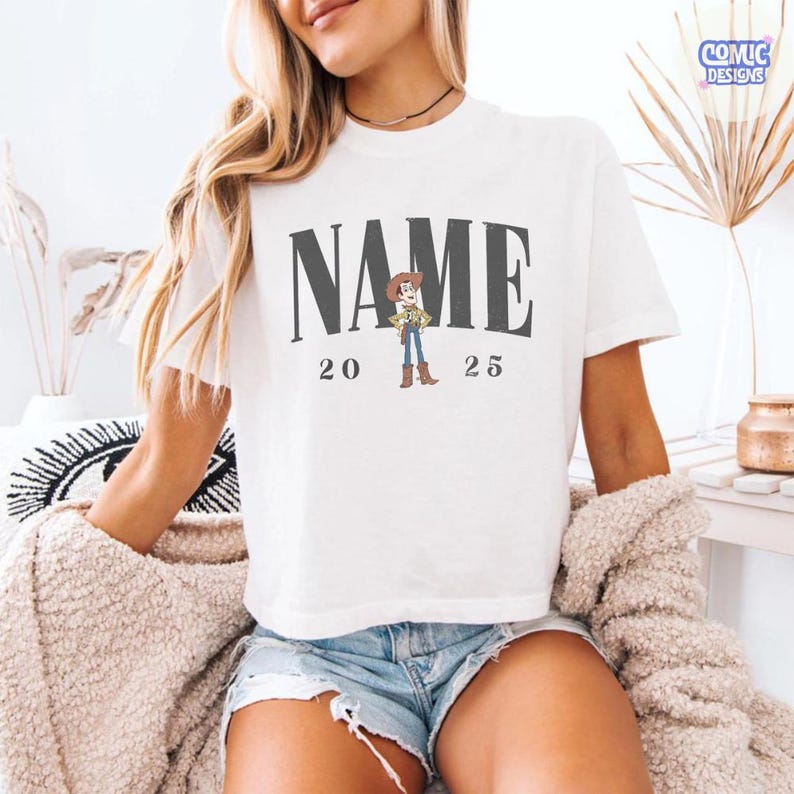 K&ouml;nnte beinhalten: Wei&szlig;es T-Shirt mit dem Wort "NAME" in dunkelgrauen Blockbuchstaben, einer Cartoon-Cowboy-Grafik und den Zahlen "20 25" darunter. Das T-Shirt hat kurze &Auml;rmel.