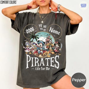 May include: Pepper-colored t-shirt featuring a pirate-themed graphic. The design includes cartoon characters, a pirate ship, and the word "PIRATES" in large letters. The text "2026" and "Life for Me" are also visible. The shirt has a relaxed fit.