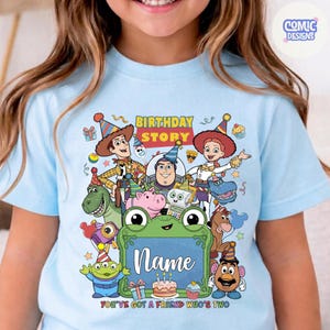 May include: A light blue t-shirt featuring a colorful graphic of Toy Story characters wearing party hats. The design includes the text "BIRTHDAY STORY" and "YOU'VE GOT A FRIEND IN ME" with a central green frog frame for a custom name.