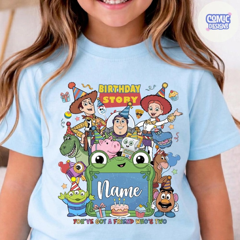 May include: A light blue t-shirt featuring a colorful graphic of Toy Story characters wearing party hats. The design includes the text "BIRTHDAY STORY" and "YOU'VE GOT A FRIEND IN ME" with a central green frog frame for a custom name.
