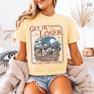 May include: Yellow t-shirt with a pirate-themed graphic featuring cartoon characters in a boat. The design includes the text "Get In Loser, We Are Going Pirating" in a vintage style. The shirt has short sleeves.