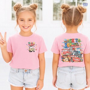 May include: A light pink short-sleeved t-shirt featuring a small graphic of a cartoon car and school supplies on the front chest, and a large colorful Back to School graphic with multiple cartoon cars on the back of the garment.