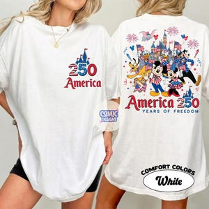 以下が含まれることがあります： 愛国的なデザインの白いTシャツ。前面には、城のグラフィックとともに、赤と青で「250 America」というテキストが表示されています。背面には、ディズニーのキャラクター、花火、旗のカラフルなイラストと、「America 250 Years of Freedom」というテキストが表示されています。