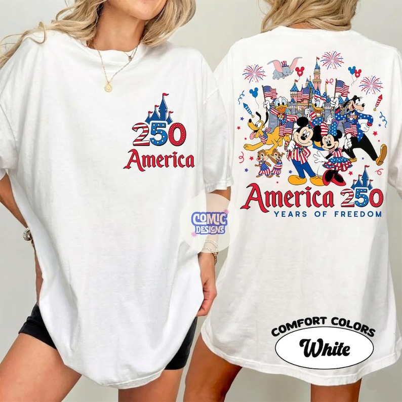 以下が含まれることがあります： 愛国的なデザインの白いTシャツ。前面には、城のグラフィックとともに、赤と青で「250 America」というテキストが表示されています。背面には、ディズニーのキャラクター、花火、旗のカラフルなイラストと、「America 250 Years of Freedom」というテキストが表示されています。