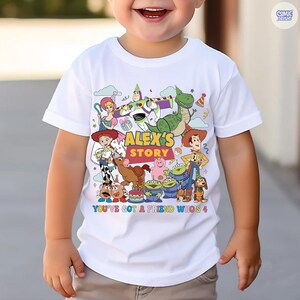 May include: White t-shirt featuring a colorful graphic of characters from the animated movie Toy Story. The shirt has the text "ALEX'S STORY" and "YOU'VE GOT A FRIEND WHO'S 4".