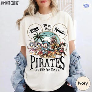 May include: Ivory-colored t-shirt with a pirate-themed graphic. The design features cartoon characters, a pirate ship, and the word "PIRATES" in large letters. Text includes "2026", "Name", and "Life for Me".