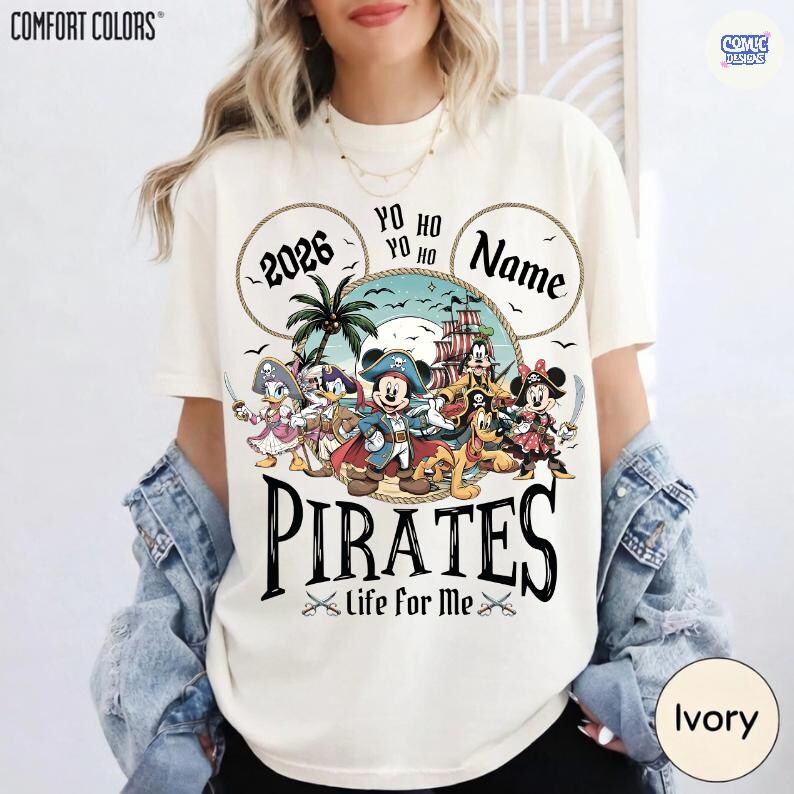 May include: Ivory-colored t-shirt with a pirate-themed graphic. The design features cartoon characters, a pirate ship, and the word "PIRATES" in large letters. Text includes "2026", "Name", and "Life for Me".