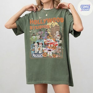 K&ouml;nnte beinhalten: Ein olivgr&uuml;nes T-Shirt mit einem Grafikdruck, der den Text "HOLLYWOOD STUDIOS" und "ROCKERS" enth&auml;lt. Das Design zeigt verschiedene Zeichentrickfiguren in Halloween-Kost&uuml;men, K&uuml;rbisse, Spinnennetze und die Silhouette eines Spukhauses.