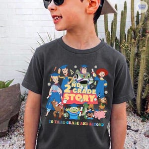 May include: Dark gray t-shirt featuring a colorful graphic of animated characters with the text "2nd Grade Story" and "To Third Grade and Beyond". The design includes characters in blue, green, red, and yellow.