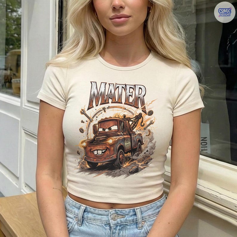 Pode incluir: Top curto creme com um gr&aacute;fico do Mate do filme Carros. O design inclui a palavra "MATER" acima do caminh&atilde;o de desenho animado, com ferramentas e um design circular ao redor do ve&iacute;culo. A camisa tem mangas curtas.