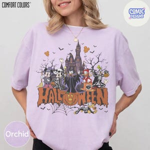 Puede incluir: Una camiseta de color morado claro con un gr&aacute;fico de personajes de Disney disfrazados de Halloween frente a un castillo. El texto HALLOWEEN est&aacute; impreso en letras grandes de color naranja en la parte inferior. Incluye a Mickey, Minnie y Goofy.