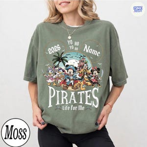 May include: Olive green t-shirt featuring a pirate-themed graphic with Mickey Mouse and friends. The design includes the text "2026", "Yo Ho Yo Ho", "Name", "Pirates", and "Life for Me".